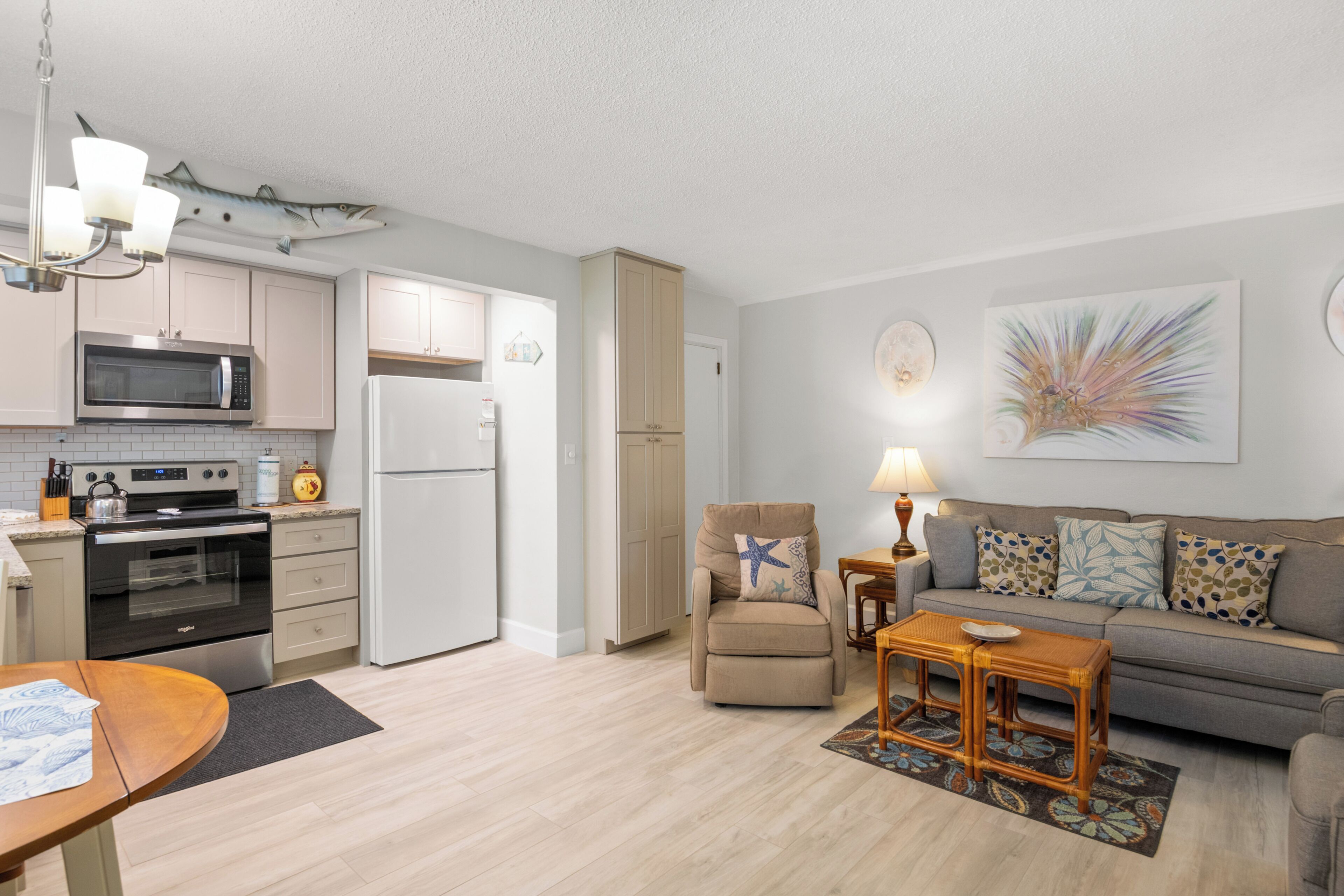 A modern kitchen with a microwave, oven, and refrigerator. There is a dining table with a placemat and a chair. A sofa and a small table with a lamp are in the living area.