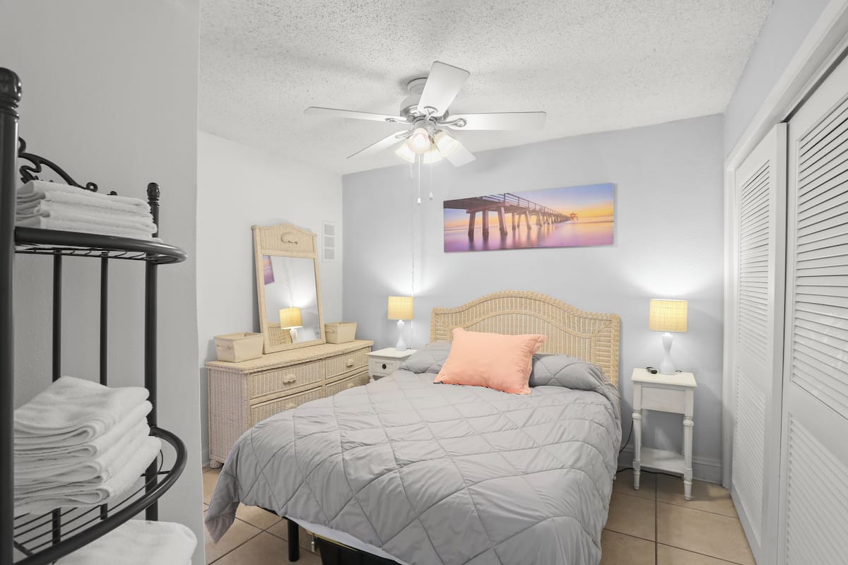 A bedroom with a bed, a ceiling fan, a nightstand with lamps, a dresser, a wall-mounted picture, and a window with blinds.