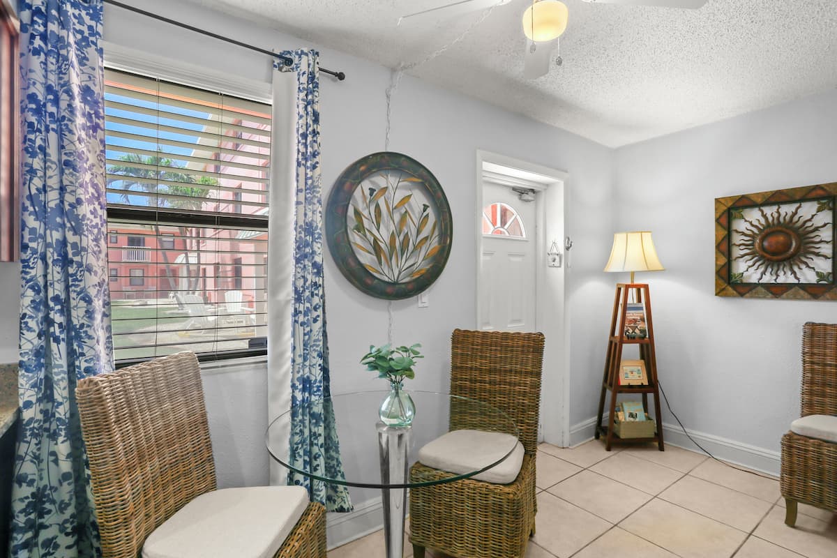 A room with wicker chairs, a glass table, a vase with a plant, a wall clock, and a sun artwork.