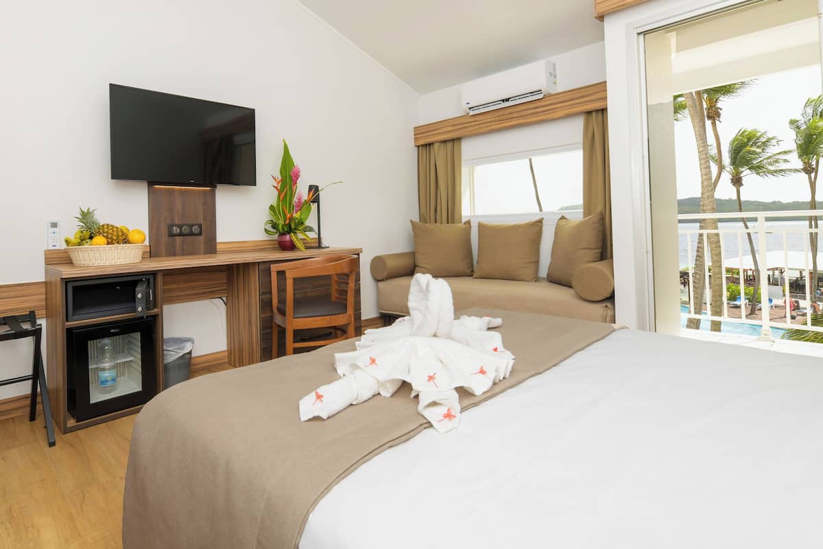 A hotel room with a bed, a desk with a TV, a small fridge, a chair, a plant, and a view of the ocean.