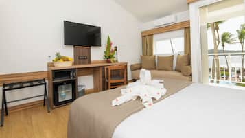 Habitación de hotel con cama, escritorio con televisor, refrigerador pequeño, silla, planta y vista al océano.