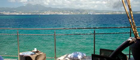 A deck with wicker furniture, a small table, and a view of the sea and distant mountains.