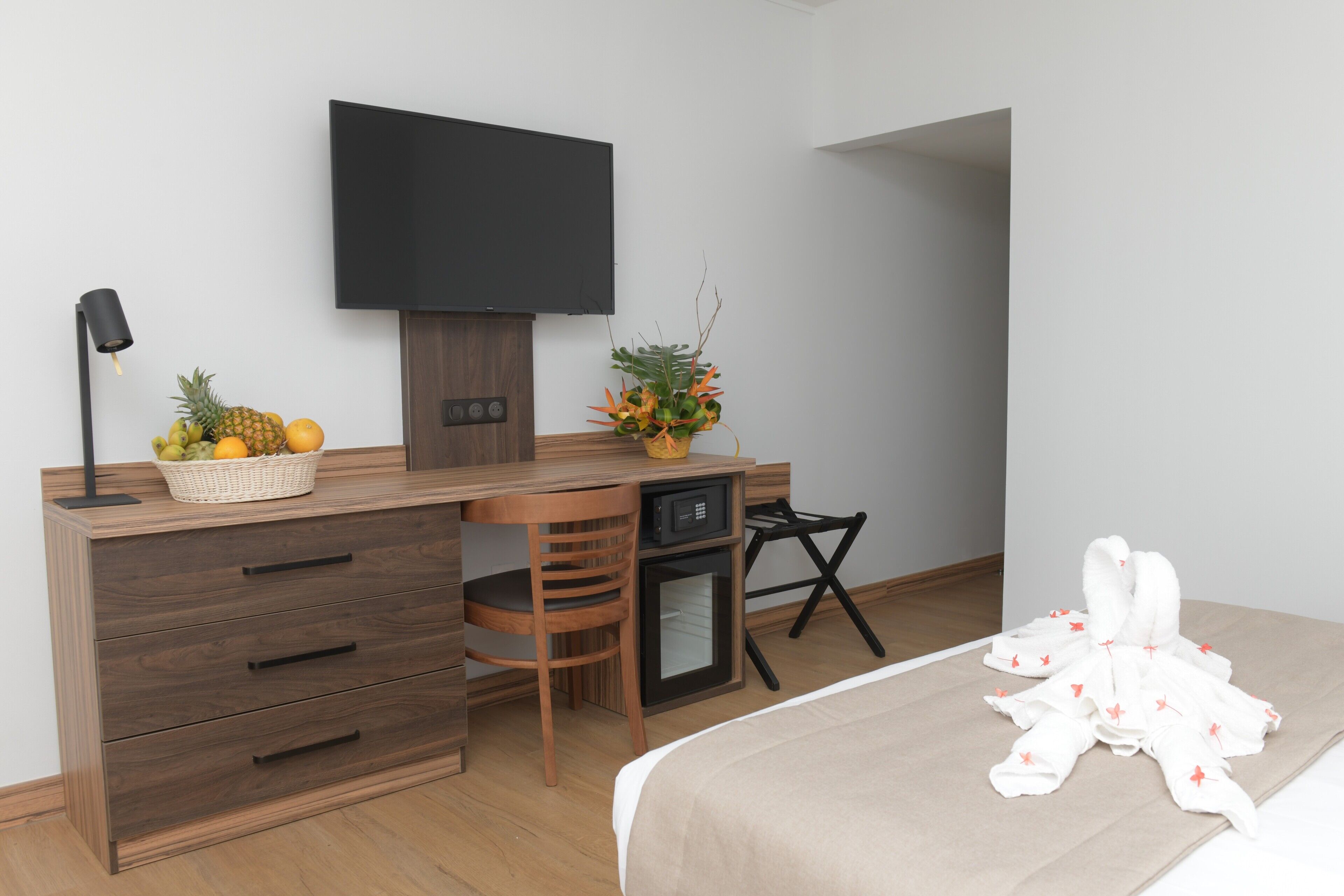 A modern hotel room with a wooden desk, a flat-screen TV, a small fridge, a chair, a bed with a white blanket, and a fruit basket.