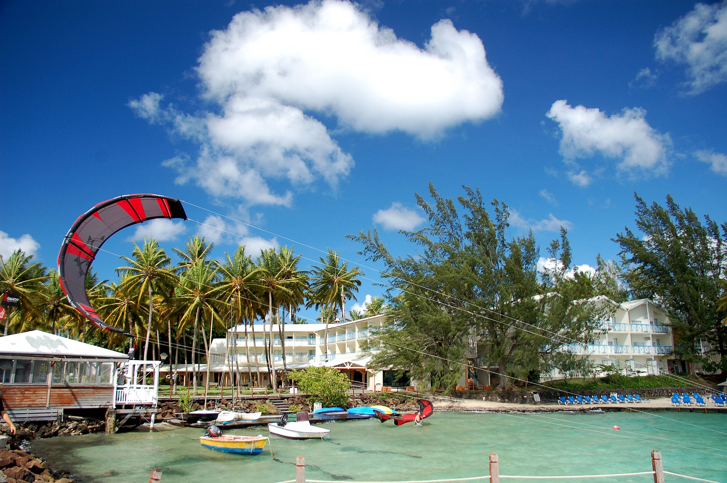 Un hotel frente a la playa con un parapente, palmeras y un cielo azul claro.
