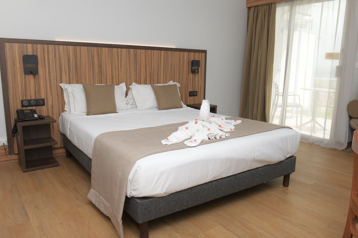 A hotel room with a large bed, wooden headboard, a bedside telephone, and a view of the outdoors through a window.