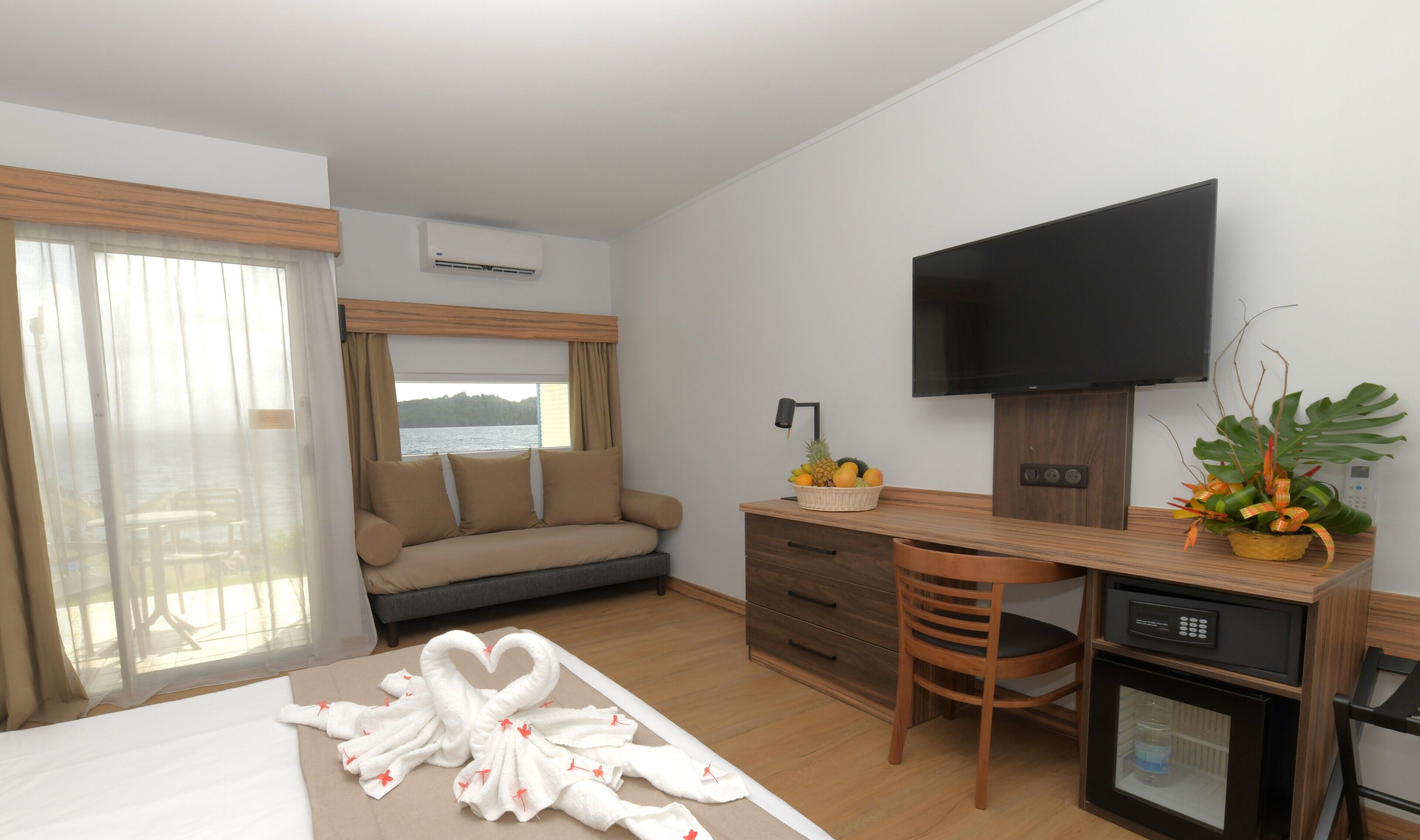 Quarto de hotel com cama, sofá, escrivaninha com televisão, pequeno frigorífico e janela com cortinas.