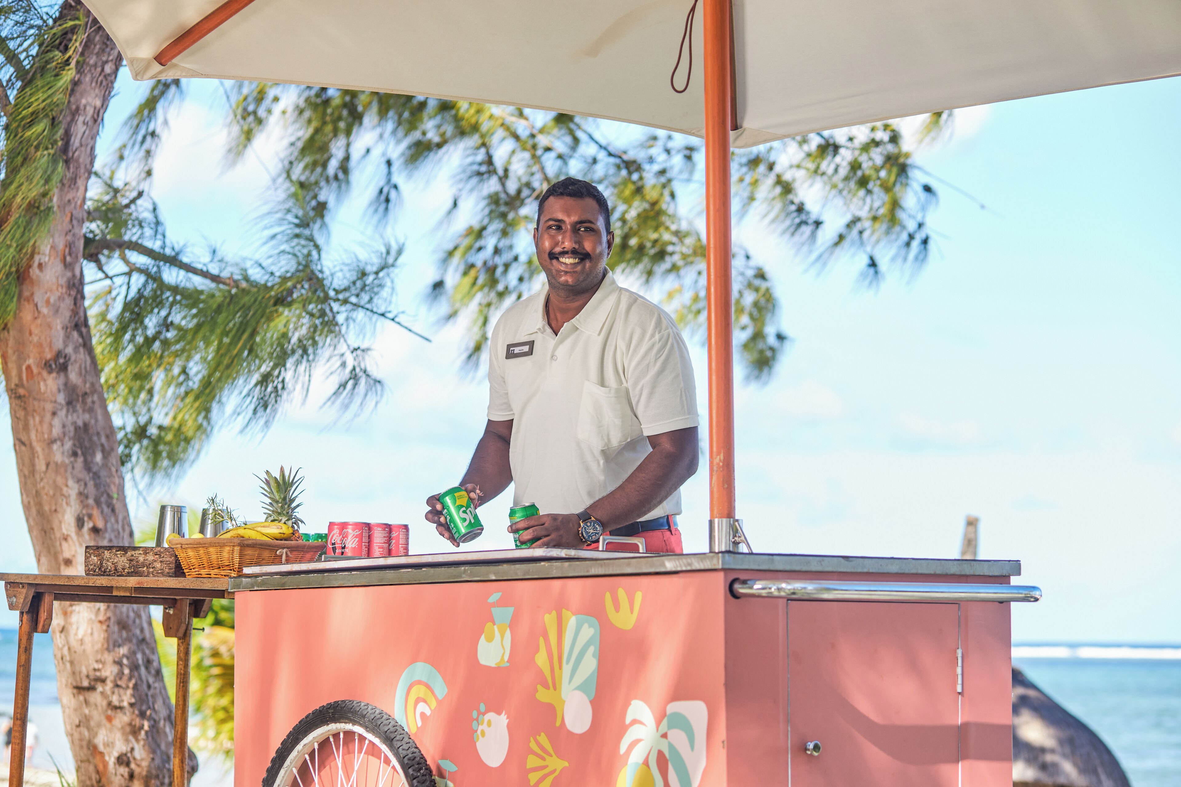 Una persona está de pie detrás de un carrito de comida con diseño tropical, cerca de una playa con palmeras y el océano al fondo.