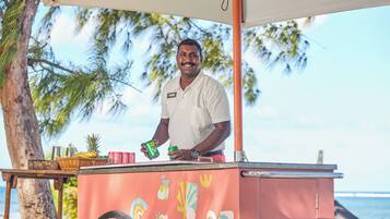 Una persona está de pie detrás de un carrito de comida con diseño tropical, cerca de una playa con palmeras y el océano al fondo.