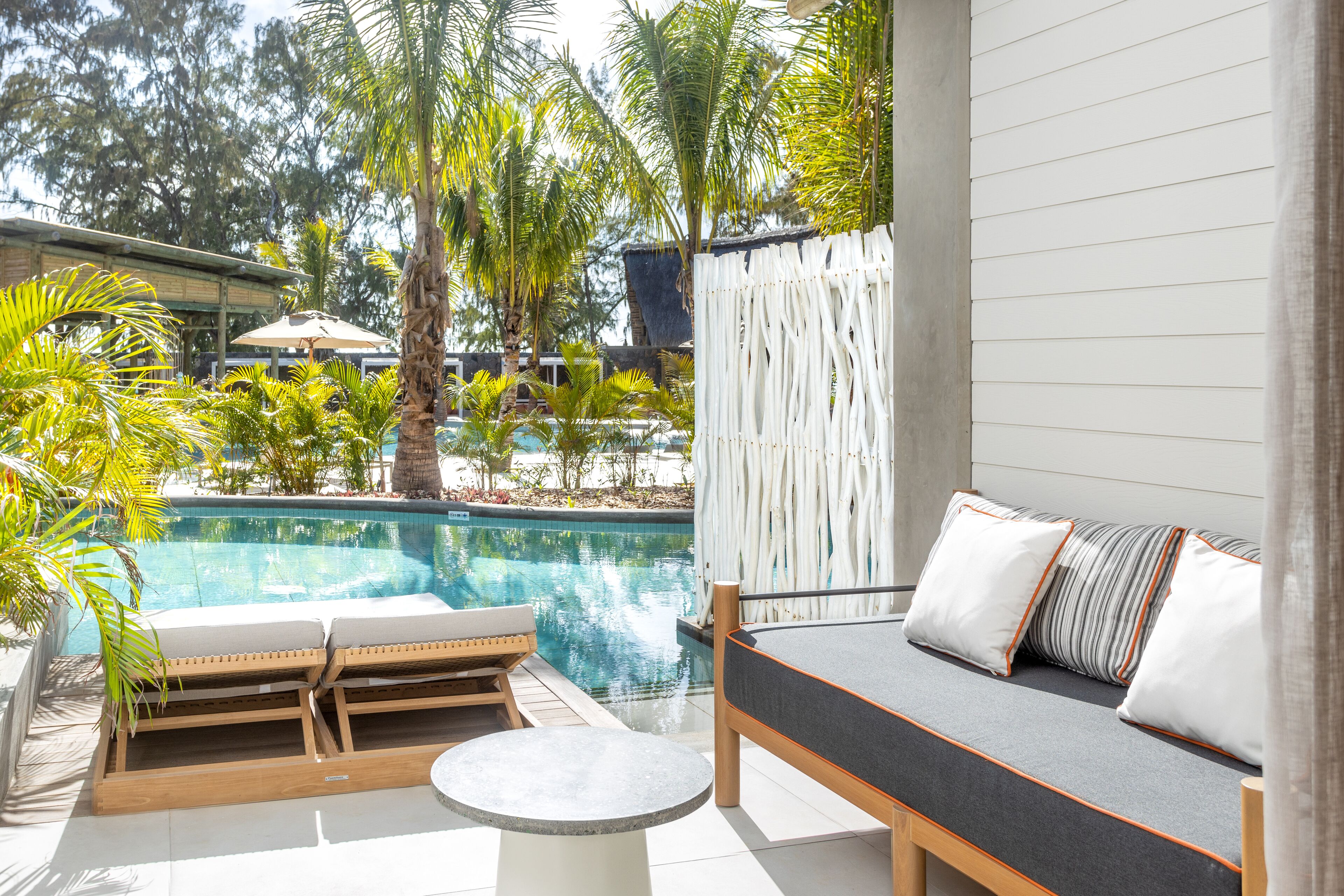 A poolside area with a wooden lounge chair, a small round table, and a sofa with cushions.