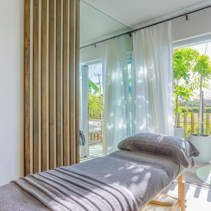 A single bed with a grey blanket and pillows, a glass door leading to an outdoor area with a wooden deck and greenery.