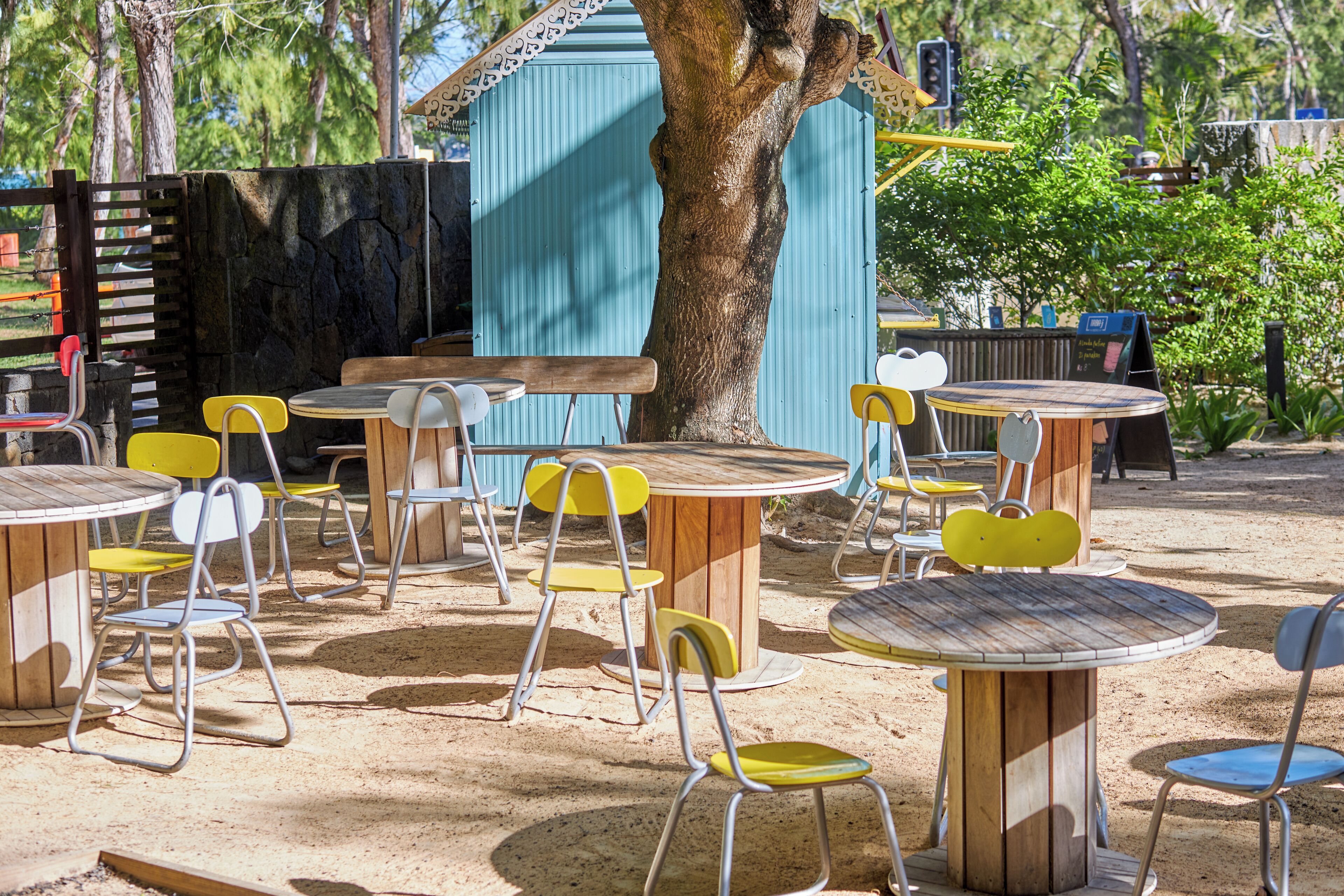 Outdoor seating with round tables and chairs, a large tree, and a blue building in the background.