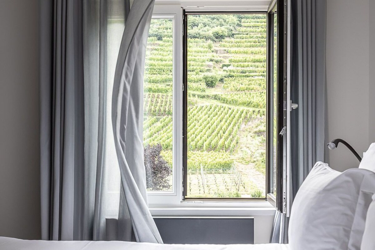 Un dormitorio con un ventanal grande, ropa de cama blanca y vistas a un viñedo.