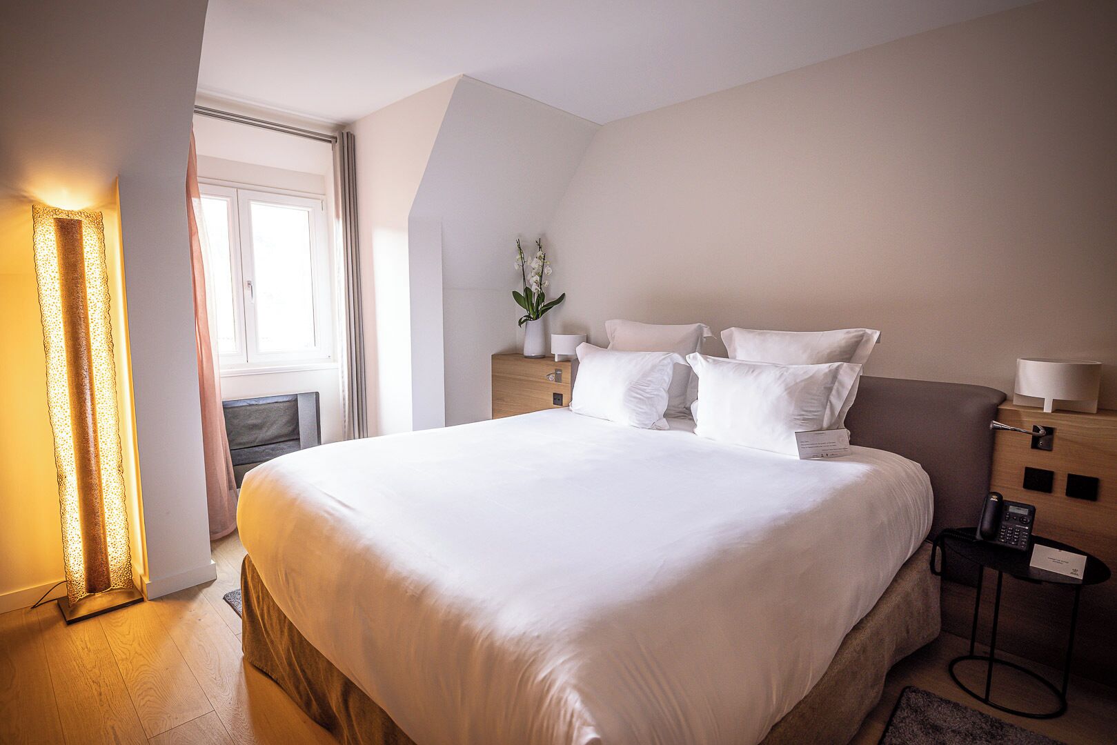 A hotel room with a large bed, bedside tables, a nightstand, a lamp, and a window.