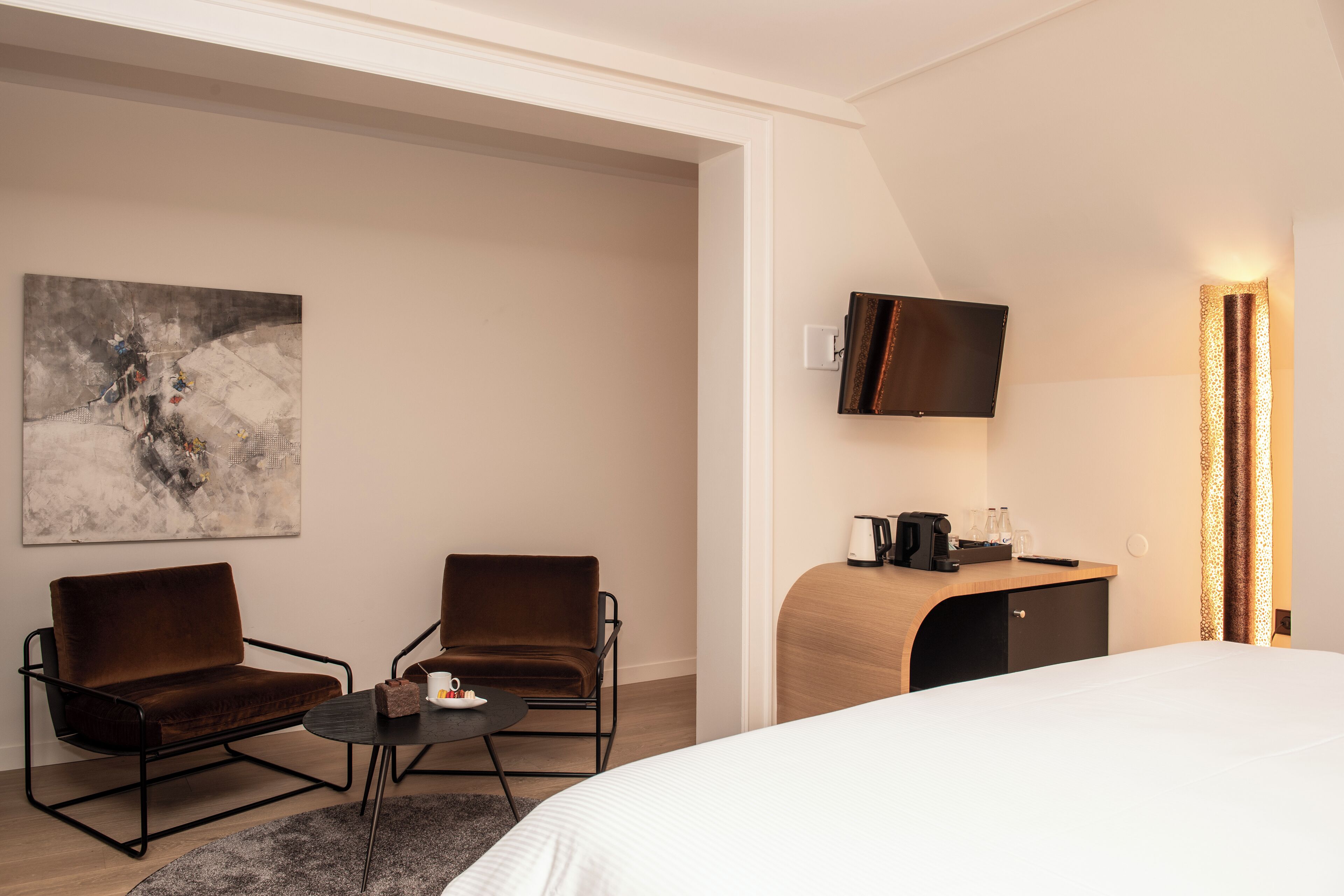 Habitación de hotel con una cama, dos sillas, una mesita, un cuadro, televisión y un escritorio con lámpara.