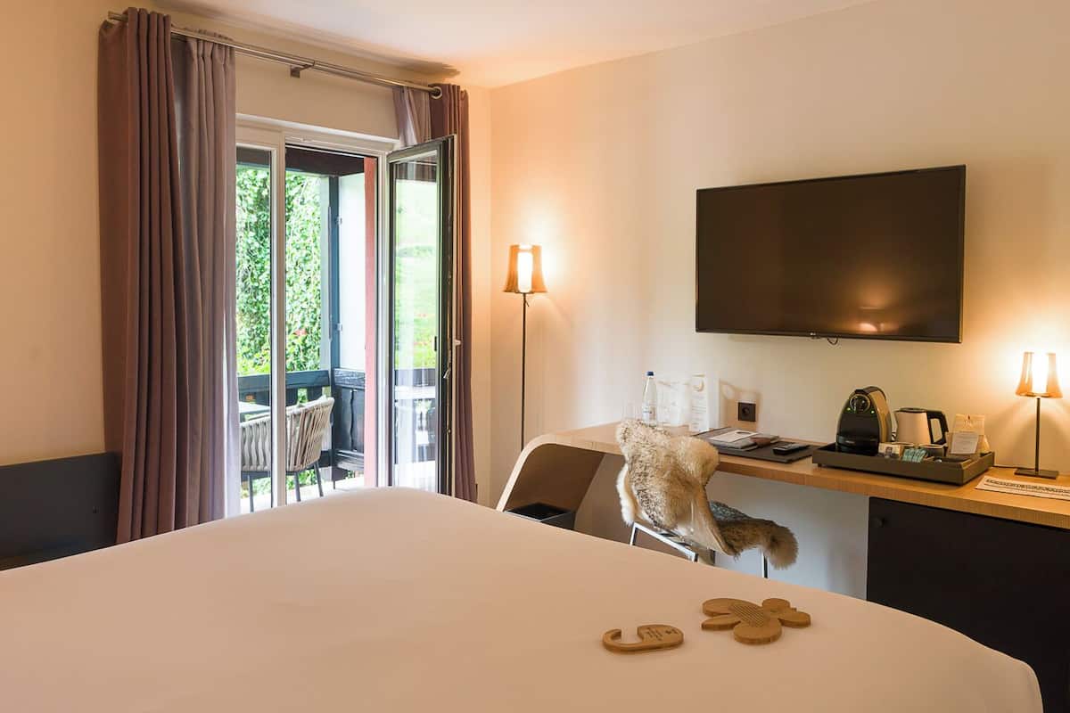 A hotel room with a bed, a desk with a chair, a television, and a balcony with curtains.