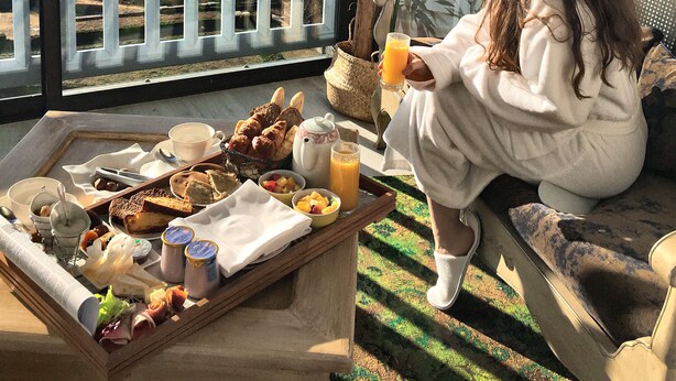 Una persona sentada junto a una ventana, con una bandeja de comida y bebidas, admirando una vista panorámica de montañas y un lago.