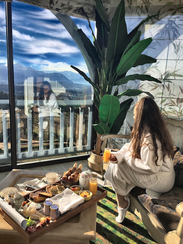 A person sitting by a window with a tray of food and drinks, overlooking a scenic view of mountains and a lake. - Château des Avenieres (Cruseilles)
