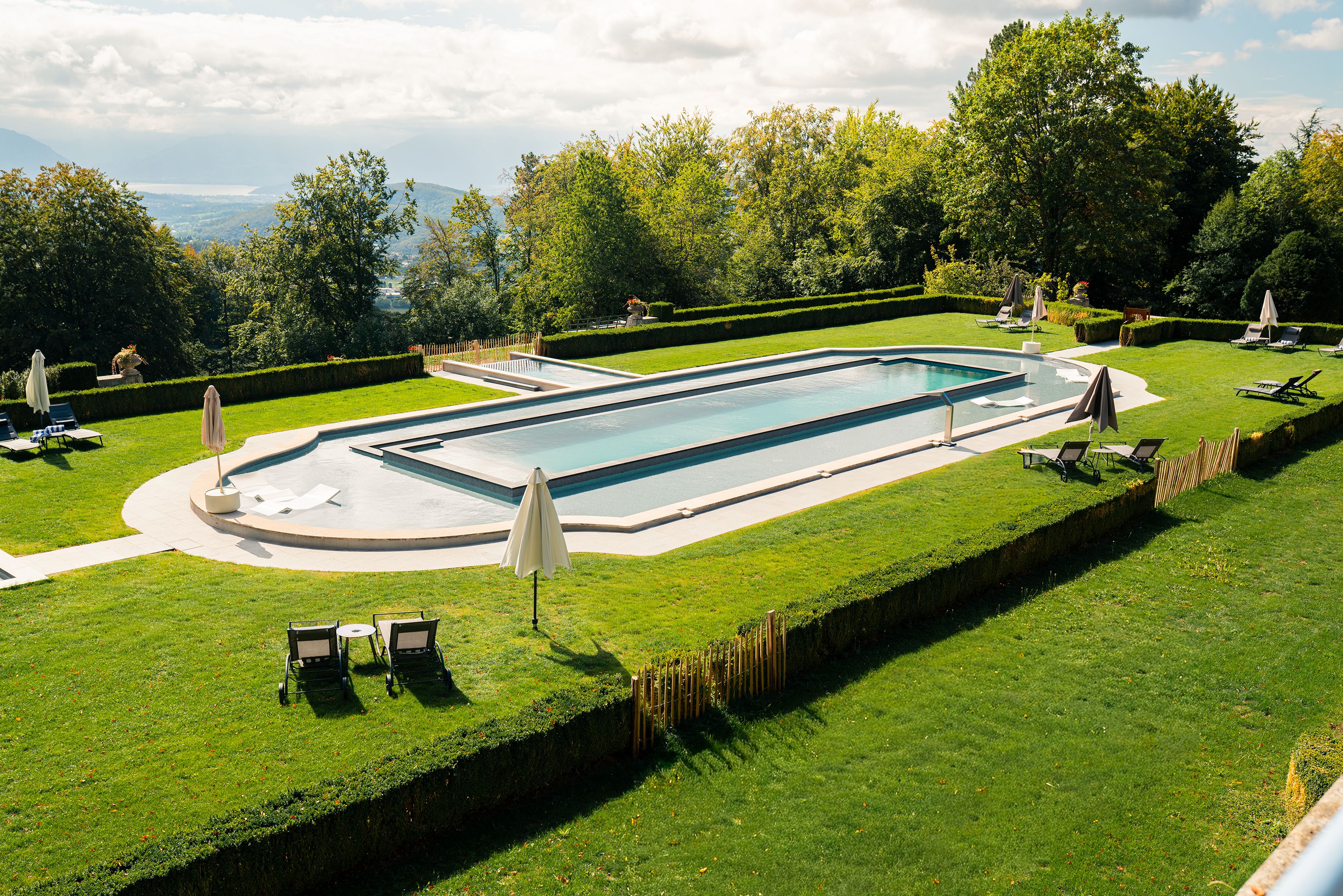 A rectangular swimming pool with a surrounding deck, surrounded by well-manicured grass and hedges, with lounge chairs and umbrellas.