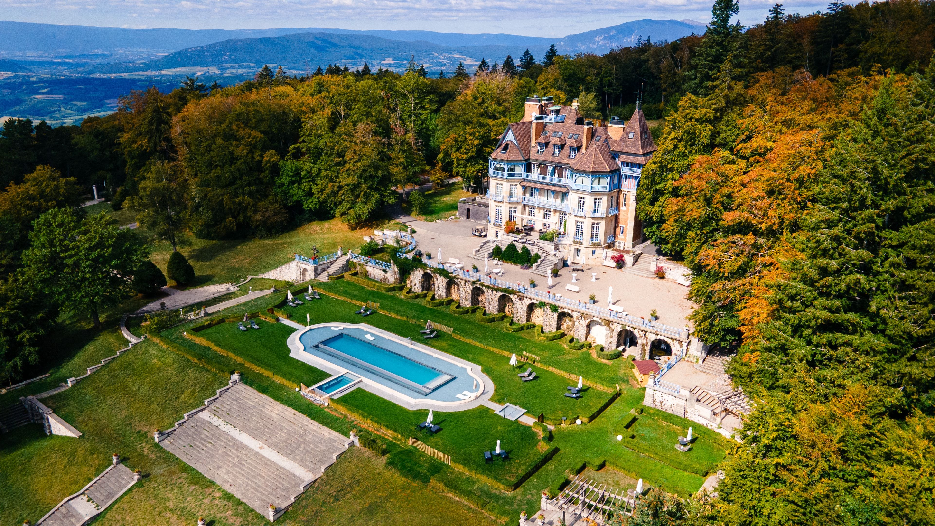 Un edificio grande e histórico con piscina, rodeado de jardines cuidados y un fondo de bosque.