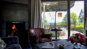Un salón con chimenea, un sillón de cuero, una mesa de centro con un periódico, un jarrón de flores y vistas a una terraza y follaje exterior.