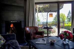 Un salón con chimenea, un sillón de cuero, una mesa de centro con un periódico, un jarrón de flores y vistas a una terraza y follaje exterior.
