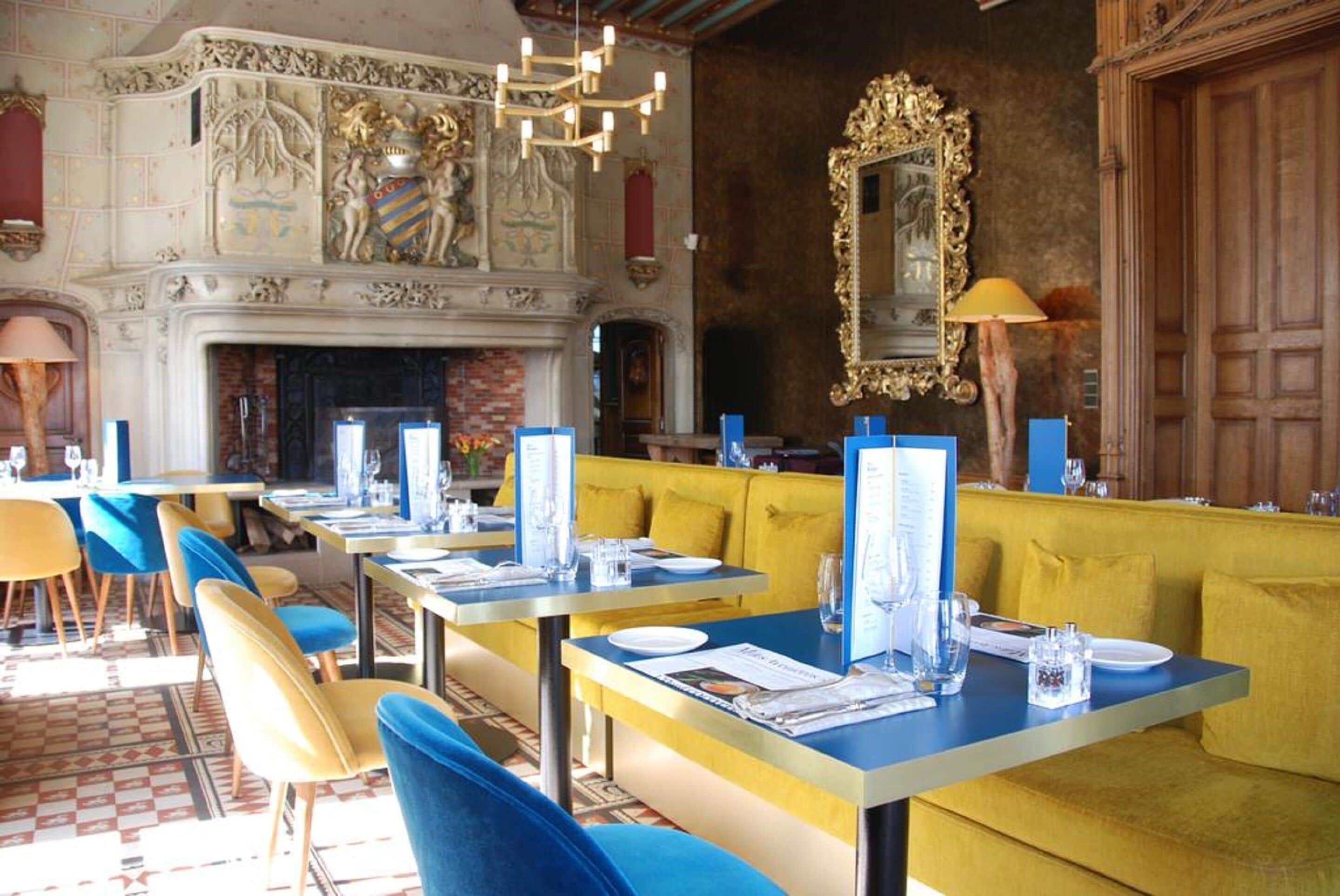 Un restaurant avec des banquettes jaunes, des chaises bleues et une cheminée. Les tables sont dressées avec des menus et des verres.