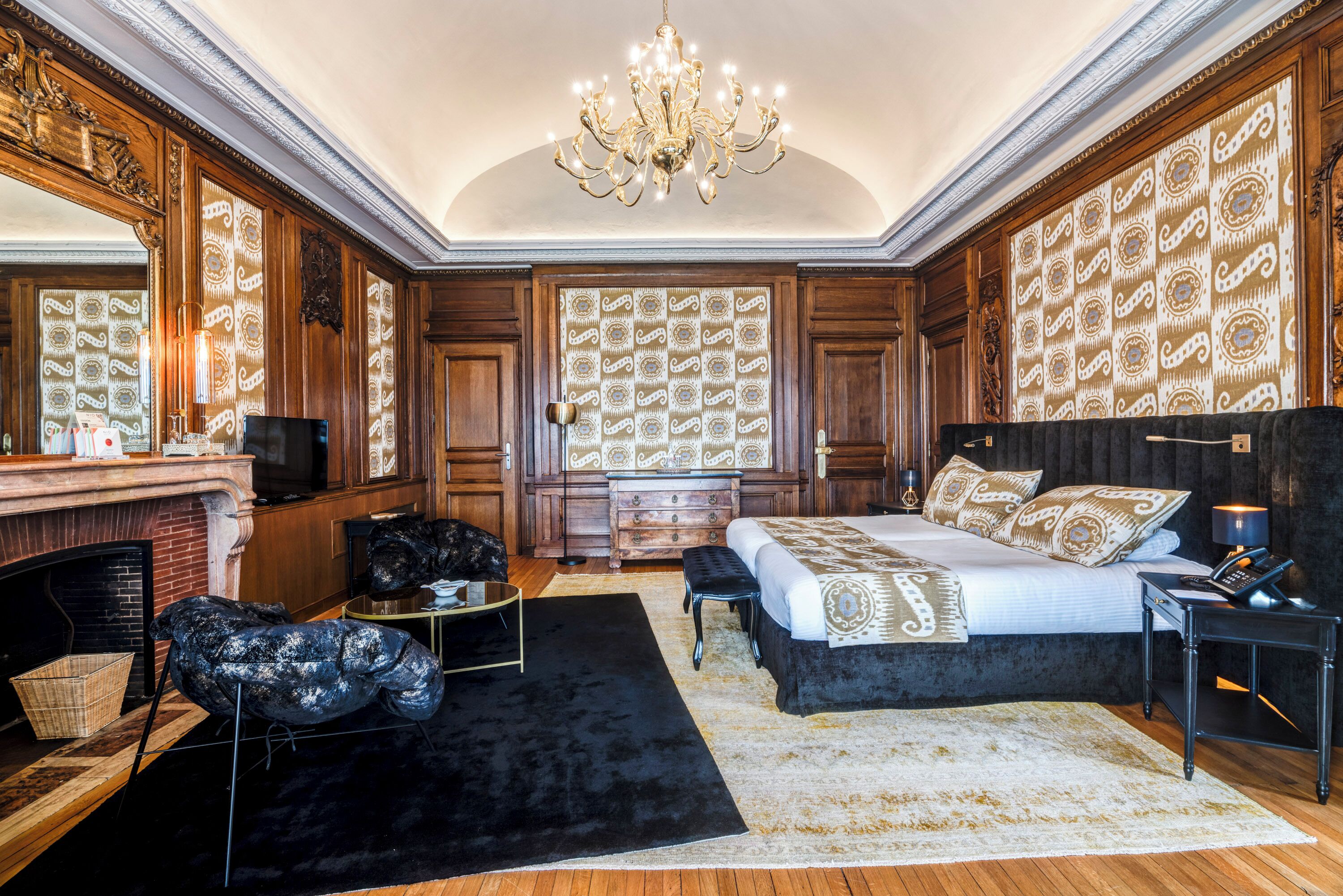 A spacious bedroom with a large bed, a fireplace, a chandelier, and ornate wallpaper.
