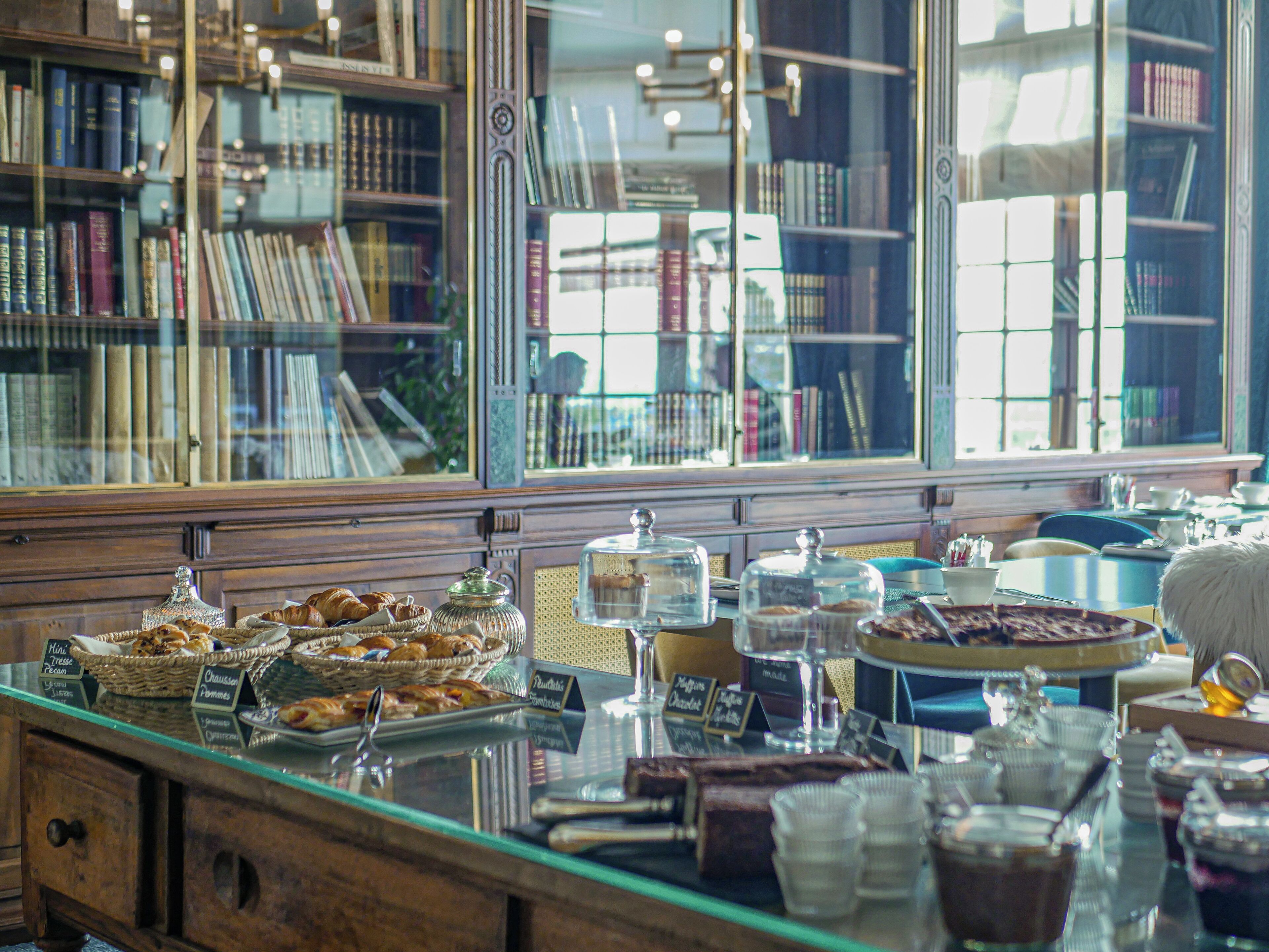 Un mostrador de panadería con diversos pasteles y postres, una vitrina de cristal y vistas a una biblioteca a través de una ventana.