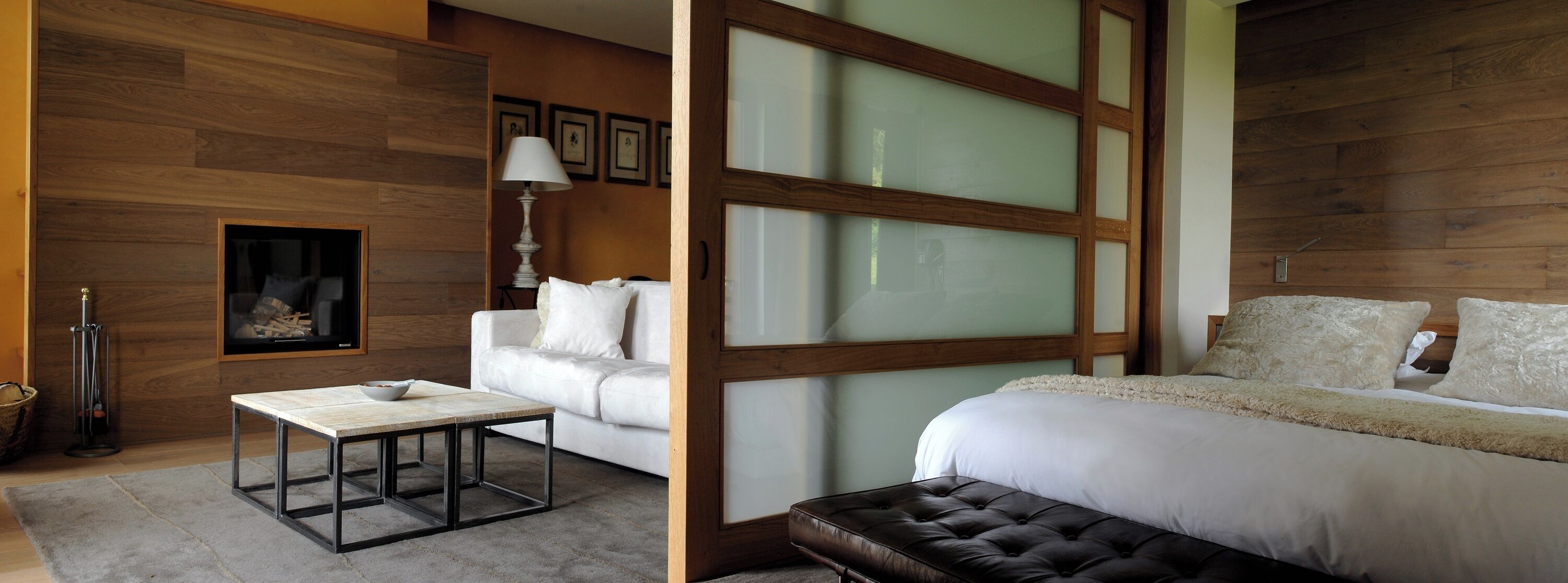 Une chambre moderne avec un plancher en bois, un canapé blanc, une cloison en verre, un lit doté d’une tête de lit capitonnée et une petite table sur laquelle se trouve un bol.