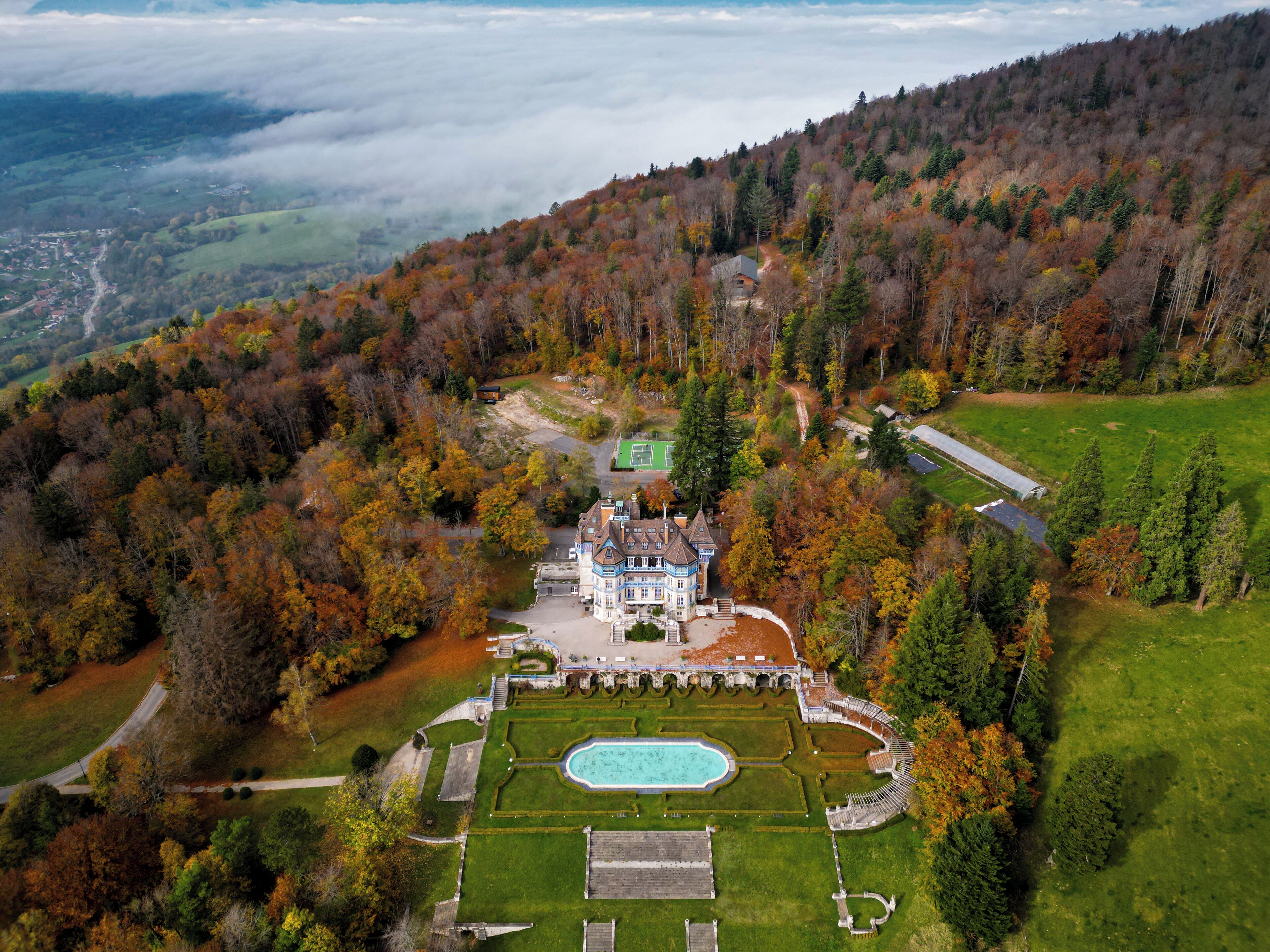 An aerial view of a large estate with a swimming pool, tennis court, and a grand house surrounded by autumn foliage.