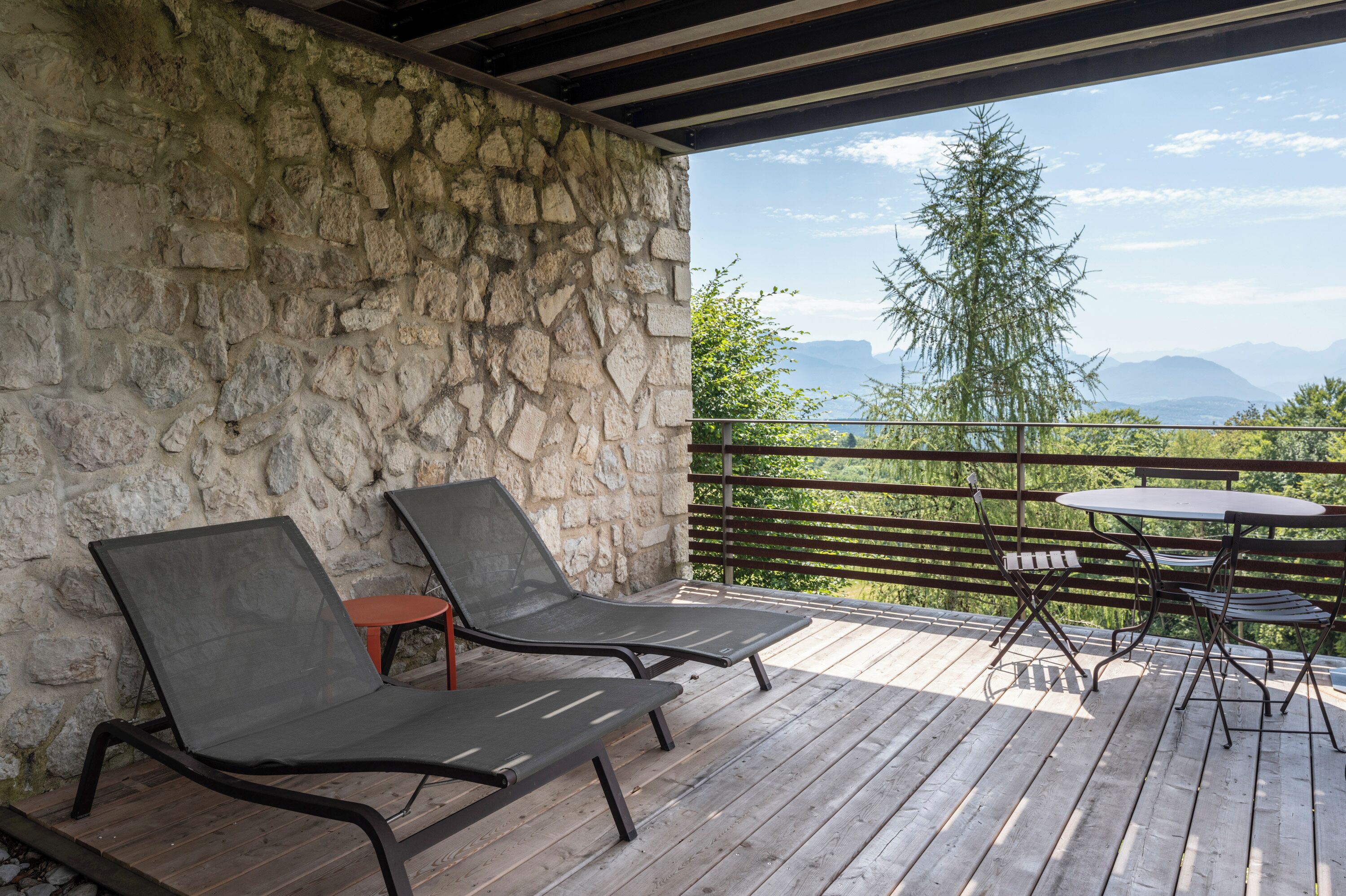 Une terrasse en bois avec deux chaises longues et une petite table, offrant une vue panoramique sur les montagnes et les arbres.