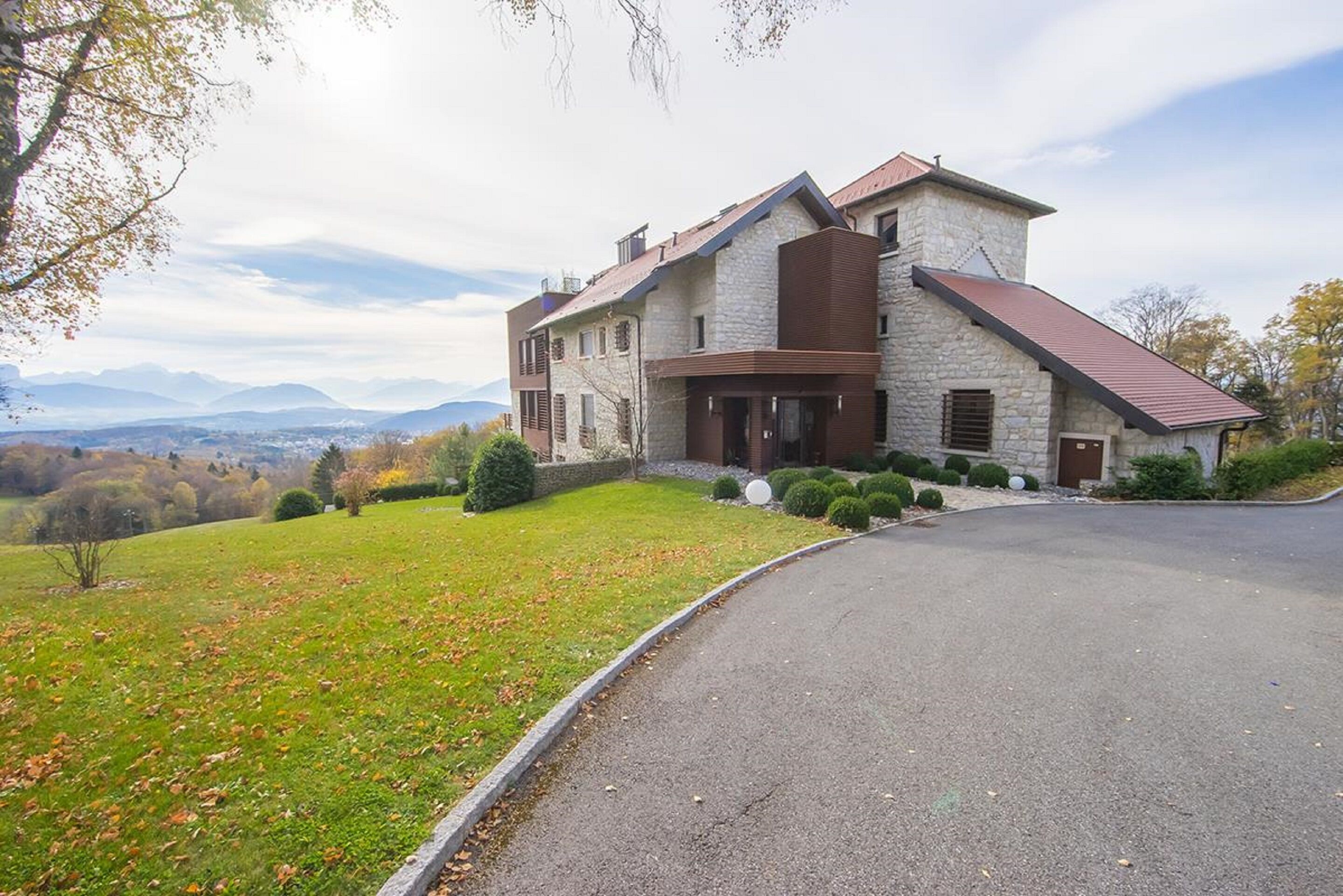 A large, modern house with a stone facade, a sloped roof, and a driveway leading to the entrance.