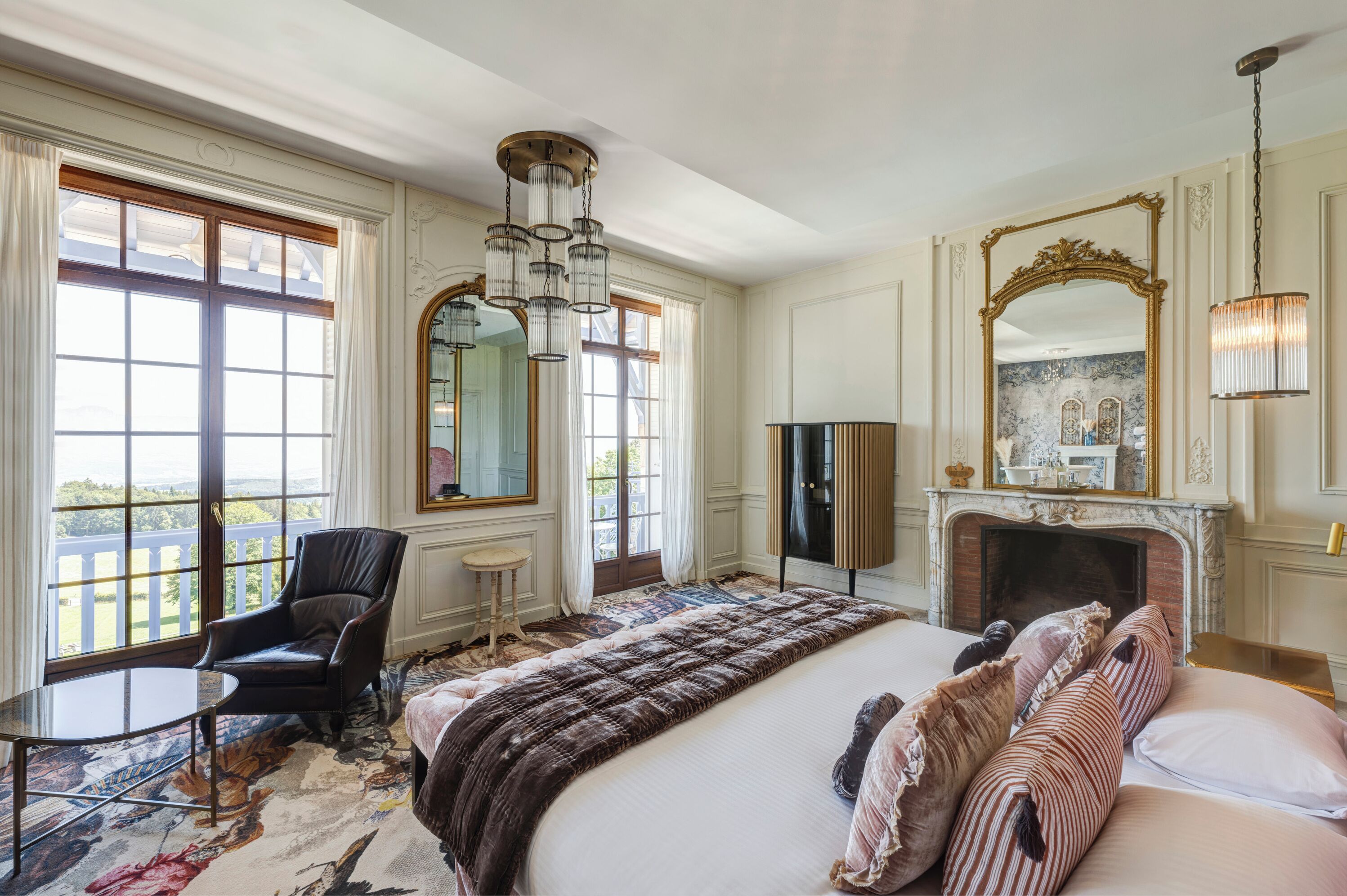 Une chambre spacieuse avec un grand lit, un coin salon avec un fauteuil, une cheminée et de grandes fenêtres offrant une vue dégagée.