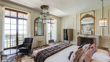 Une chambre spacieuse avec un grand lit, un coin salon avec un fauteuil, une cheminée et de grandes fenêtres offrant une vue dégagée.