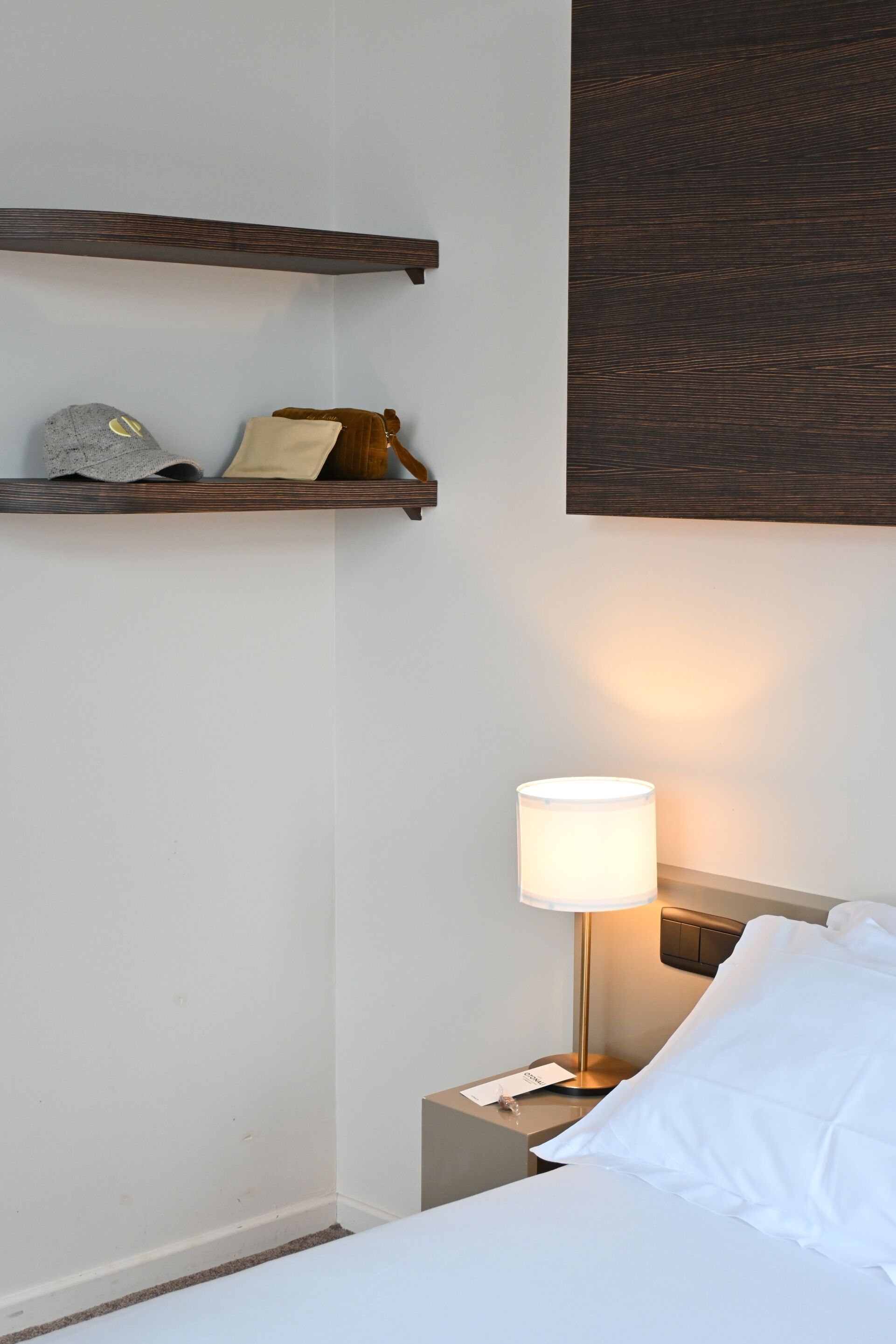 A hotel room with a bed, a nightstand with a lamp, a wooden shelf with a hat and a bag, and a dark textured wall above the shelf.