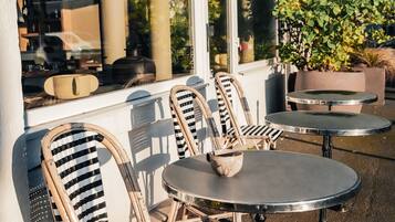 Outdoor seating with wicker chairs and round tables, a potted plant, and a building with a sign.