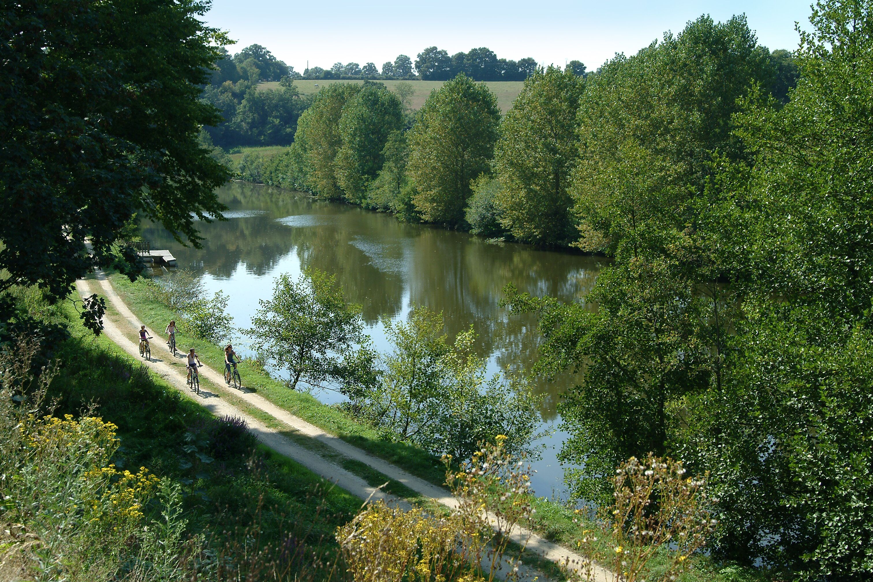 A river with cyclists on a path, surrounded by greenery.