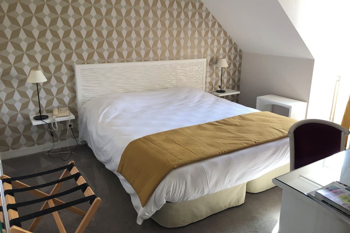 A hotel room with a bed, bedside tables, a desk, a chair, and a patterned wall.