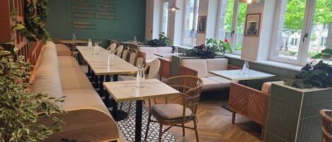 A restaurant with long communal tables, cushioned seating, and large windows.