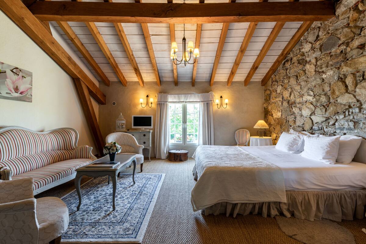 Une chambre avec un mur en pierre, des poutres apparentes, un lit, un canapé, un bureau et un lustre.