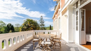 Een terras met een ronde tafel en stoelen, een witte balustrade en uitzicht op begroeiing en bergen.