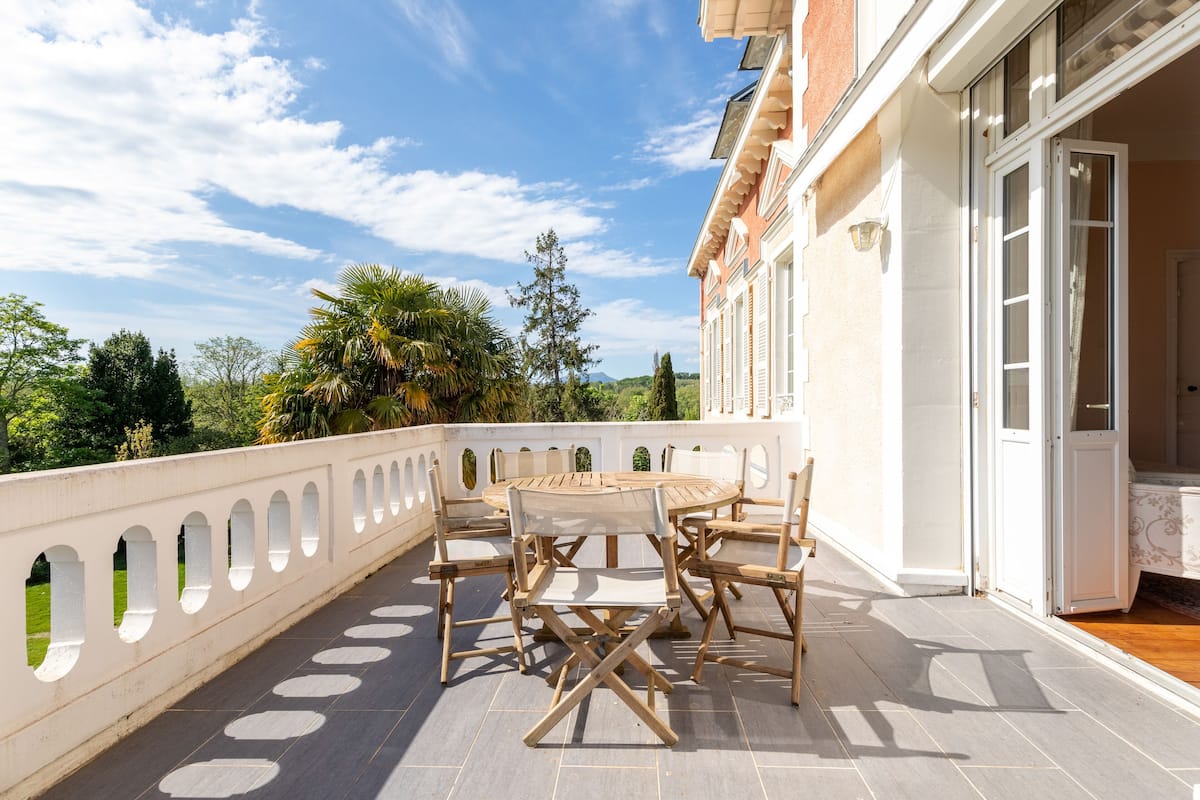 Une terrasse avec une table ronde et des chaises, une balustrade blanche et une vue sur la verdure et les montagnes.