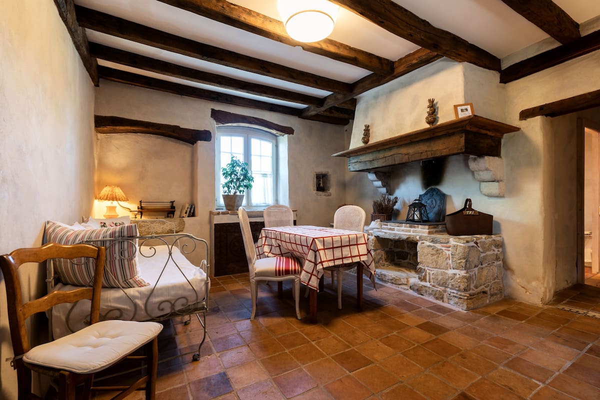 Une pièce rustique avec des poutres apparentes, une cheminée en pierre, une table à manger avec des chaises, un lit avec une tête de lit en bois et une fenêtre agrémentée d’une plante.