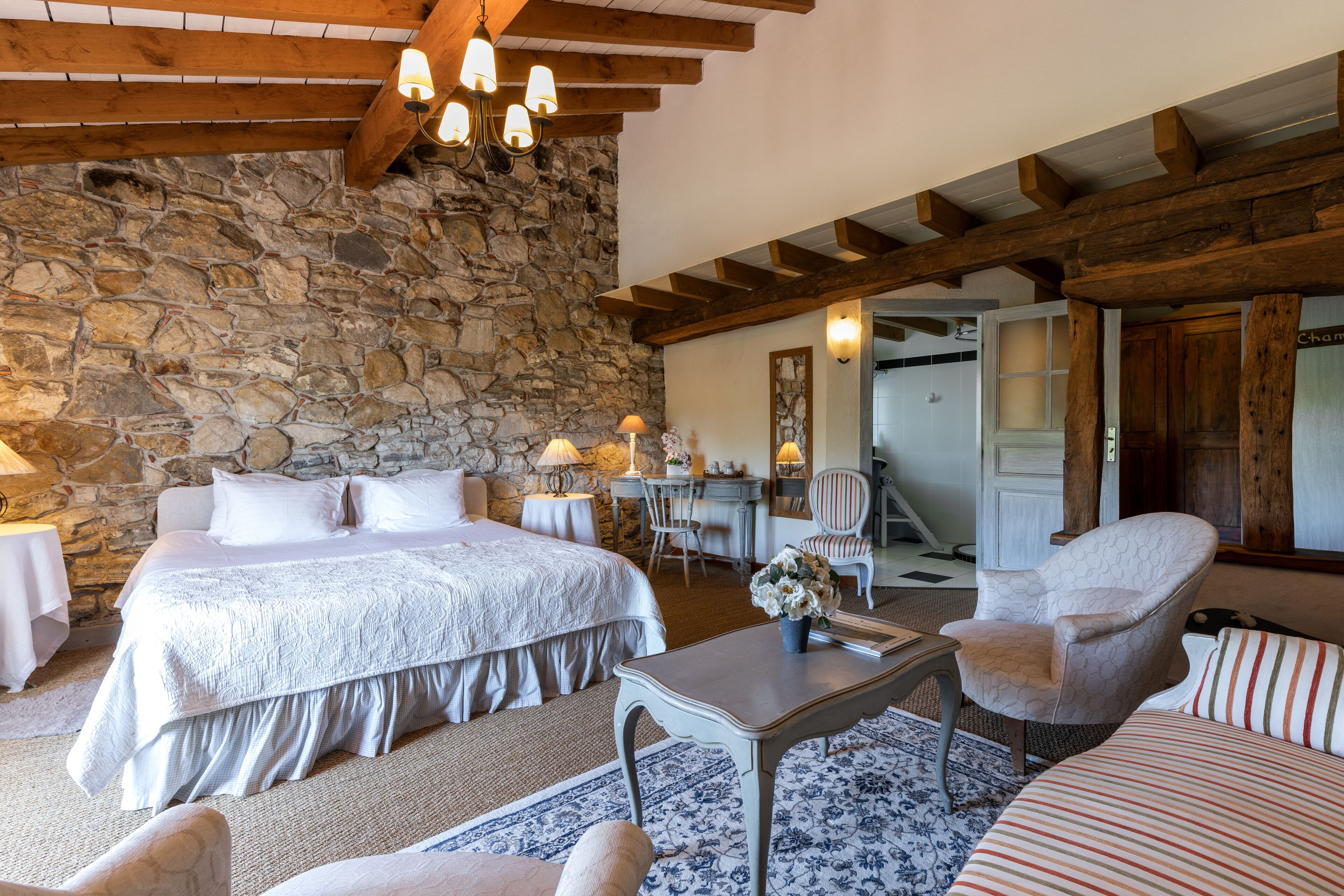 A bedroom with a stone wall, wooden beams, a large bed, a desk, and a chair.