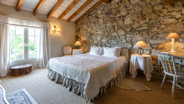 Een slaapkamer met een stenen muur, houten balken, een groot bed, twee nachtkastjes met lampen, een stoel en een raam met gordijnen.