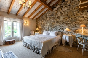 Un dormitorio con pared de piedra, vigas de madera, una cama grande, dos mesitas de noche con lámparas, una silla y una ventana con cortinas.