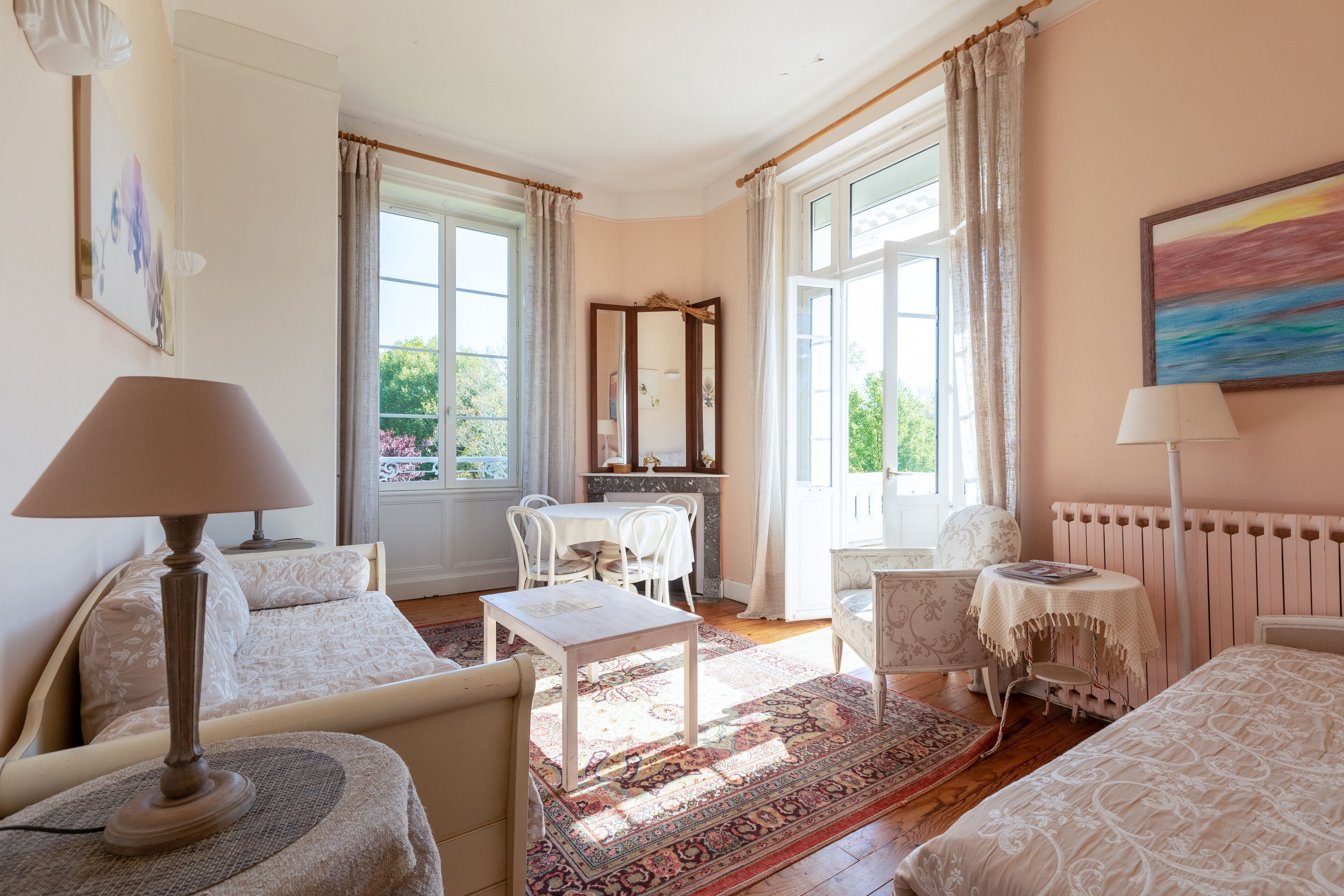 A well-lit room with a dining table, chairs, a small table with a lamp, a bed, and a large window with curtains.