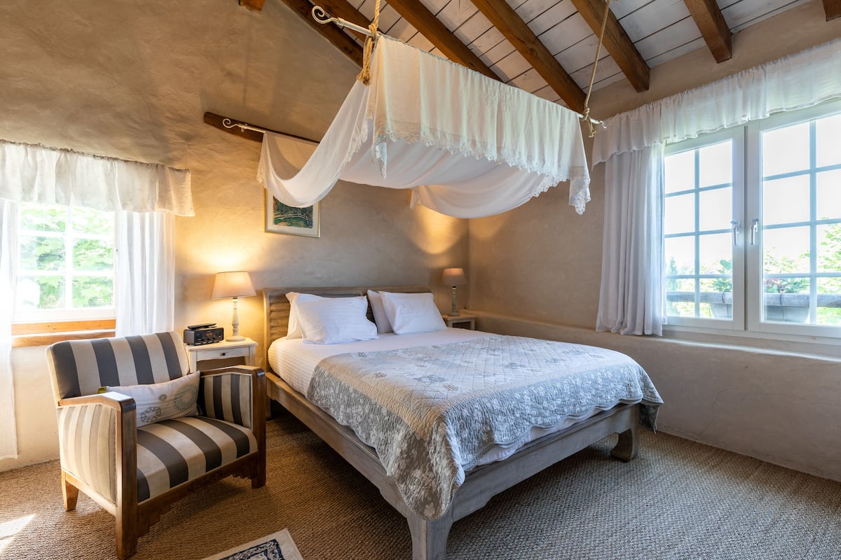 Une chambre à coucher avec un lit à baldaquin, un fauteuil à rayures, une table de chevet avec une lampe et une fenêtre avec des rideaux.