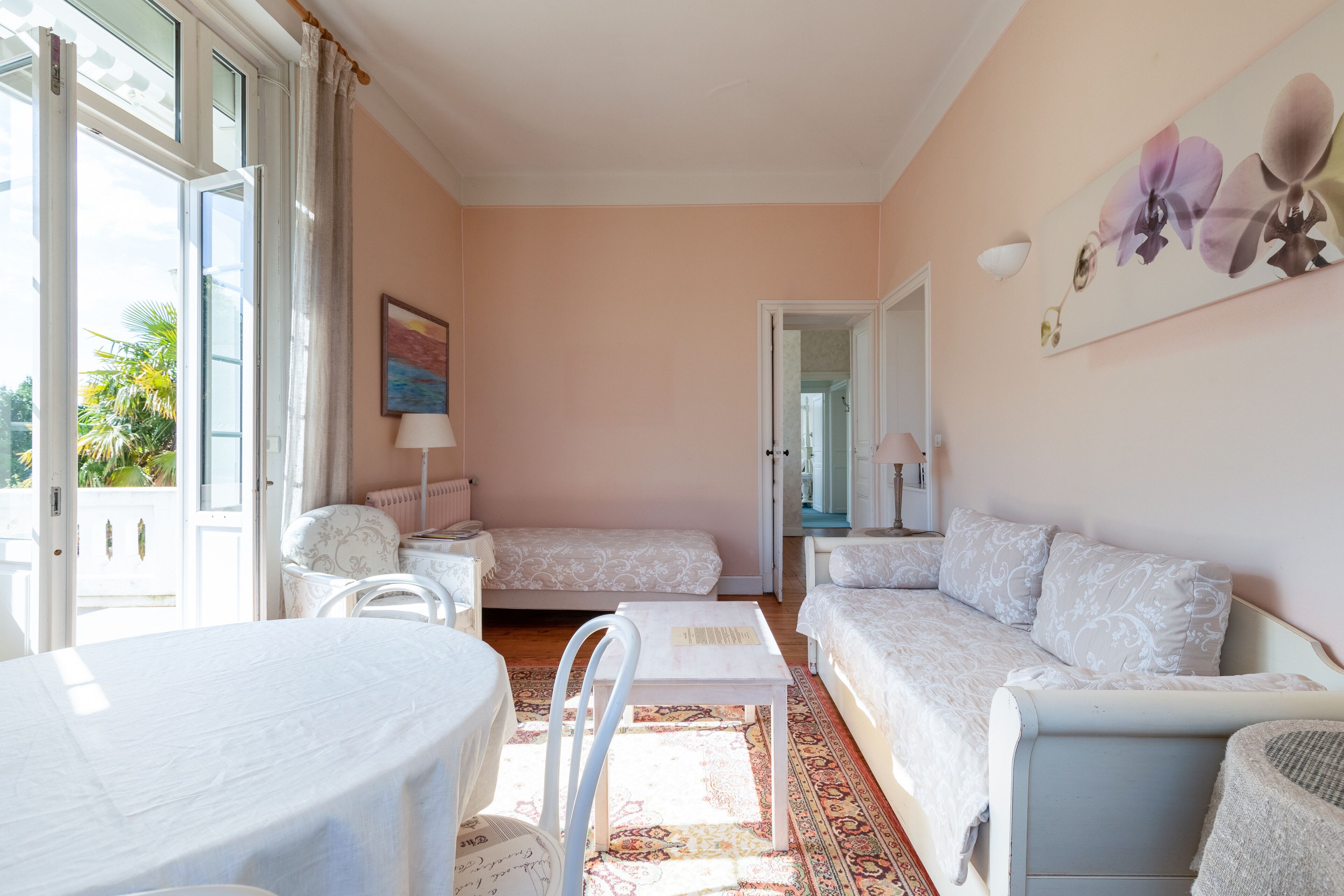 A room with a bed, a sofa, a table, and a chair. There is a window with curtains, a painting on the wall, and a door leading to another room.