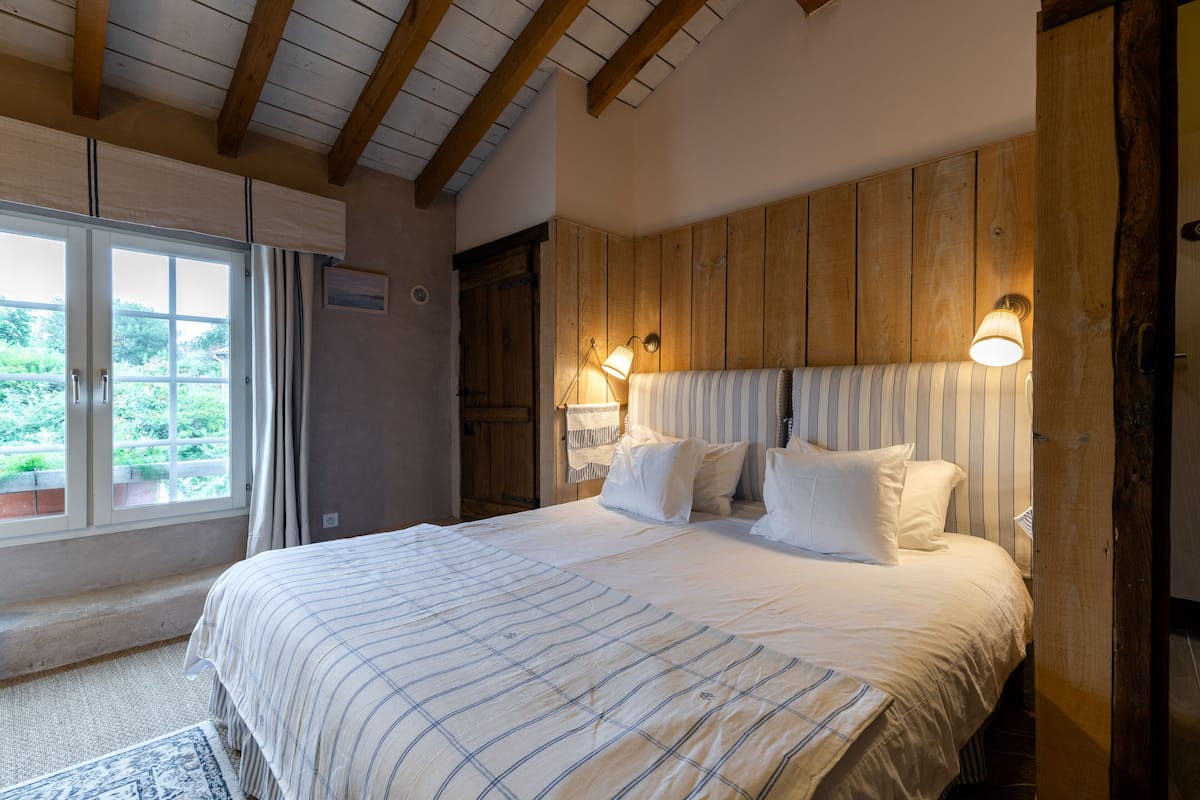 Une chambre à coucher avec un grand lit, une tête de lit en bois, deux lampes de chevet et une vue sur la verdure par la fenêtre.